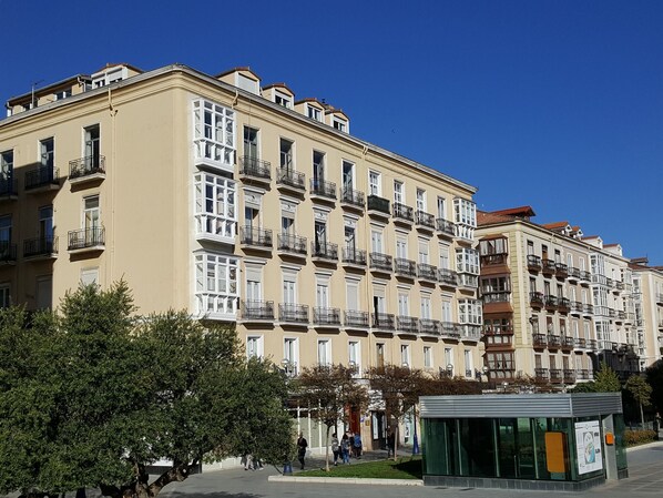 Un edificio de varios pisos con balcones, una estructura de fachada de vidrio y un cielo azul claro.