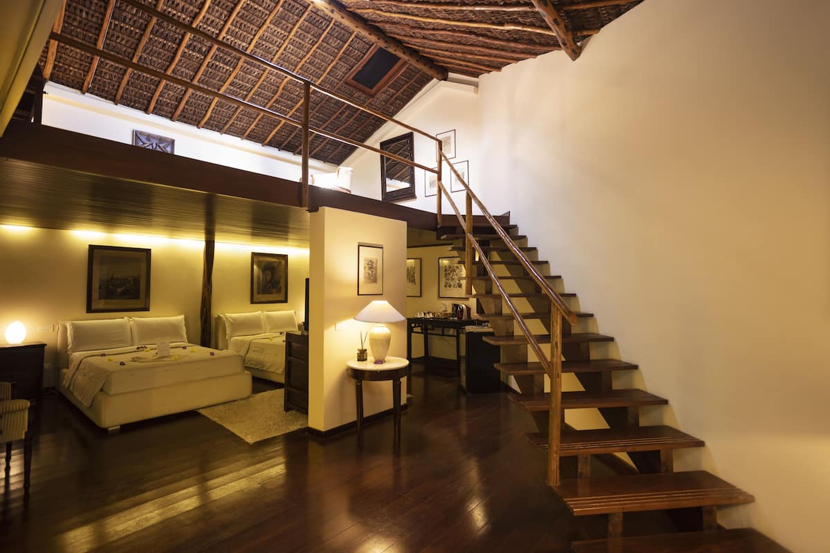 Un vaste espace de vie agrémenté d’un escalier en bois, d’un plafond haut avec une structure de poutres apparentes, et d’un coin salon confortable avec une lampe.