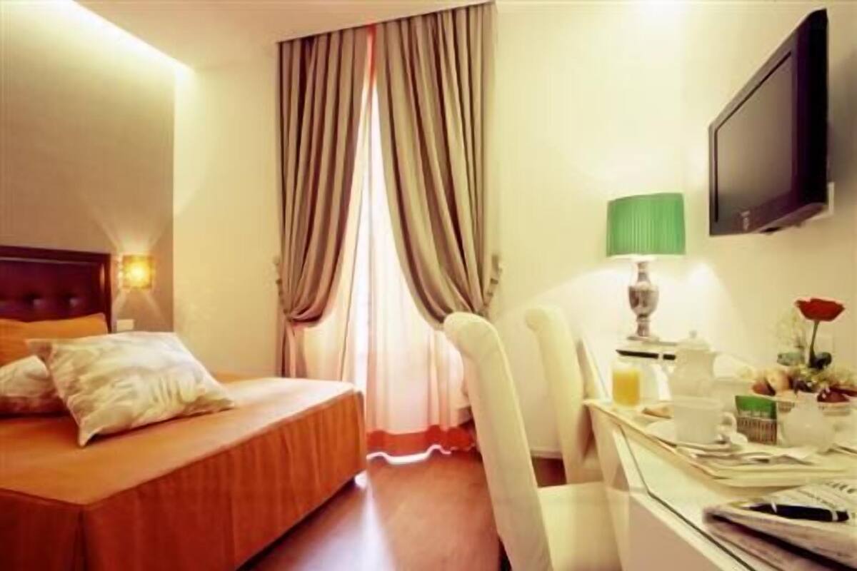 A hotel room with a bed, a desk with a lamp, a television, and a window with curtains.