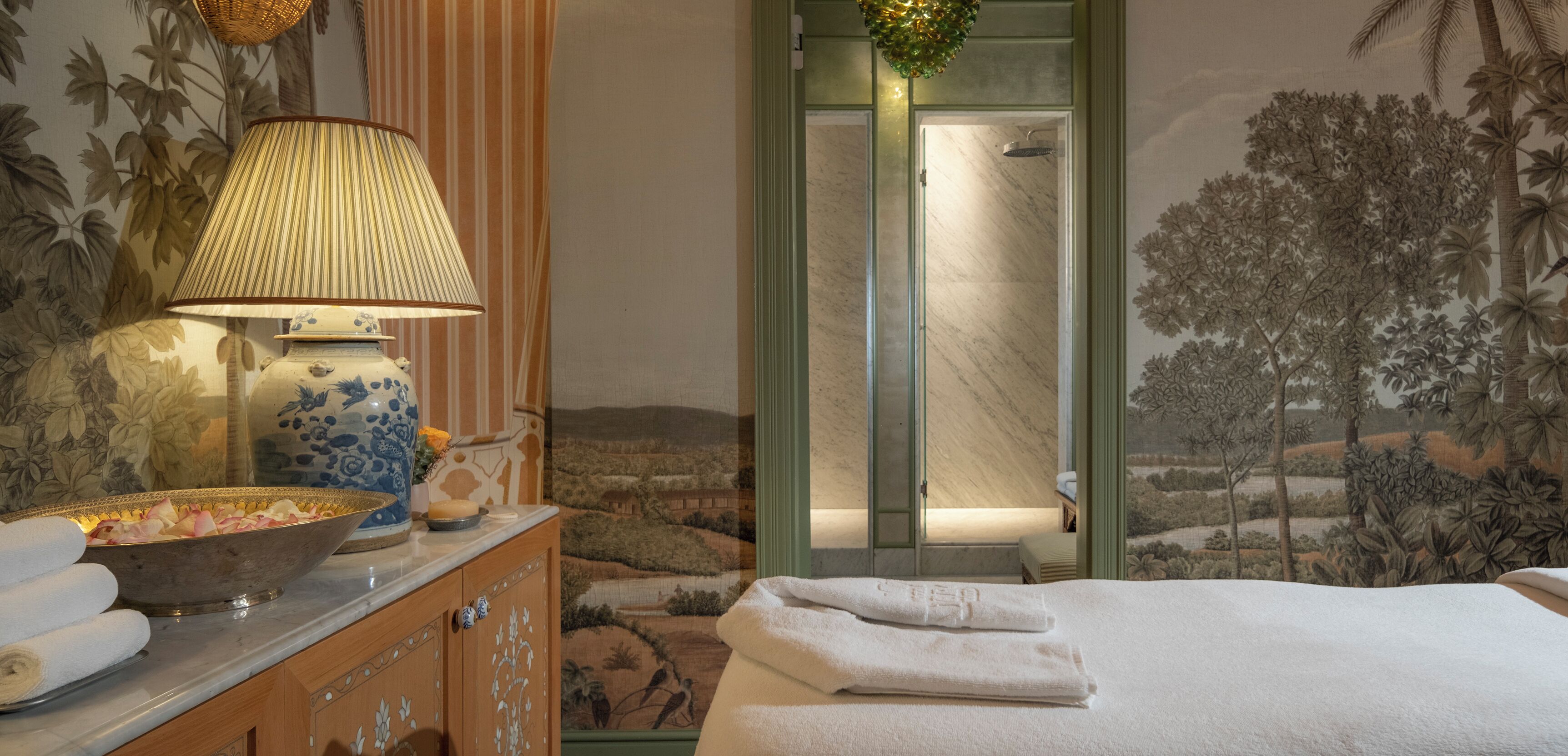 Une chambre d’hôtel avec un lit, une salle de bain avec douche, une lampe décorative et un panier de fruits posé sur la commode.