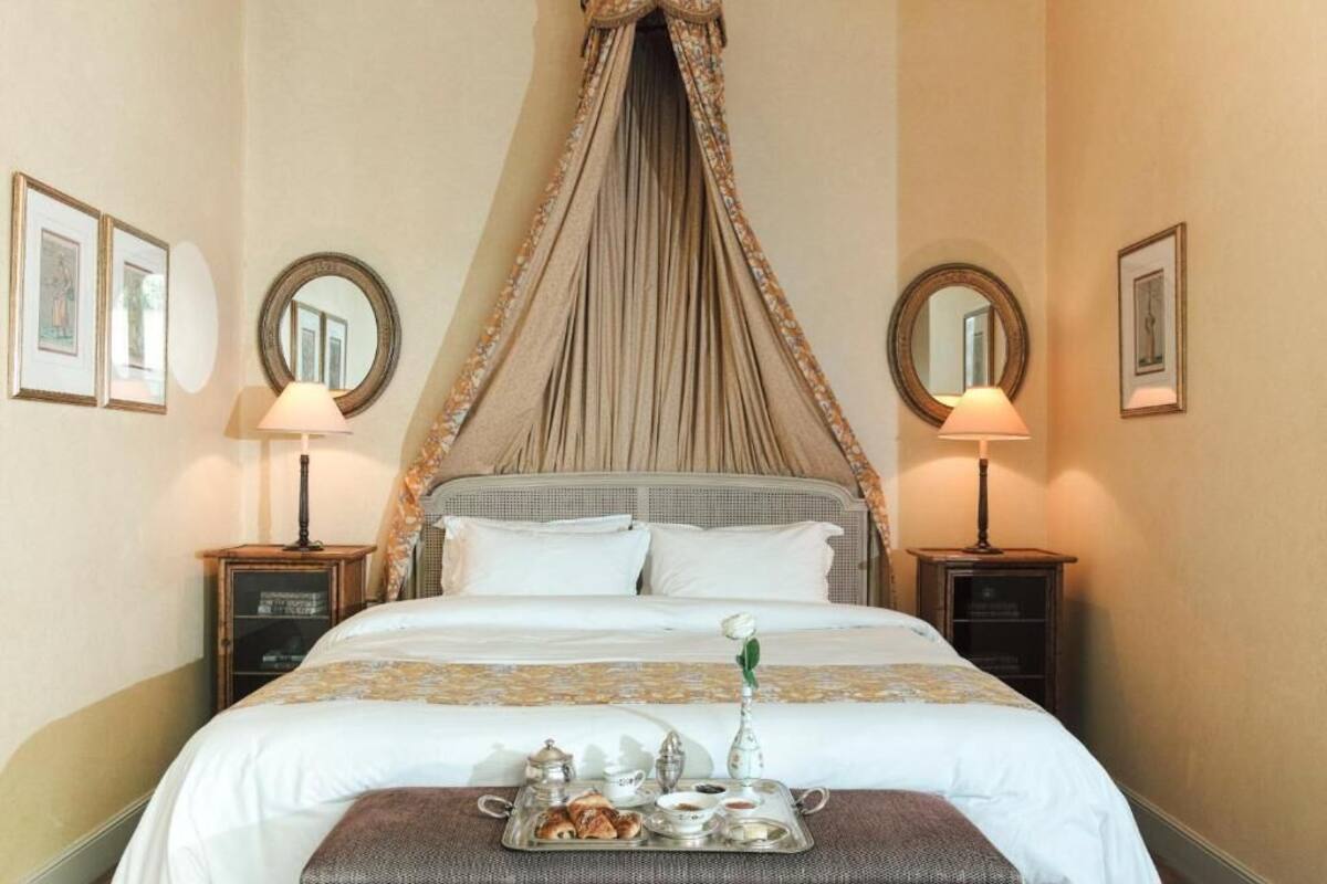 Un lit bien fait, surmonté d’un dais, deux tables de chevet avec des lampes, et un plateau contenant divers éléments pour le petit-déjeuner, posé sur le lit.