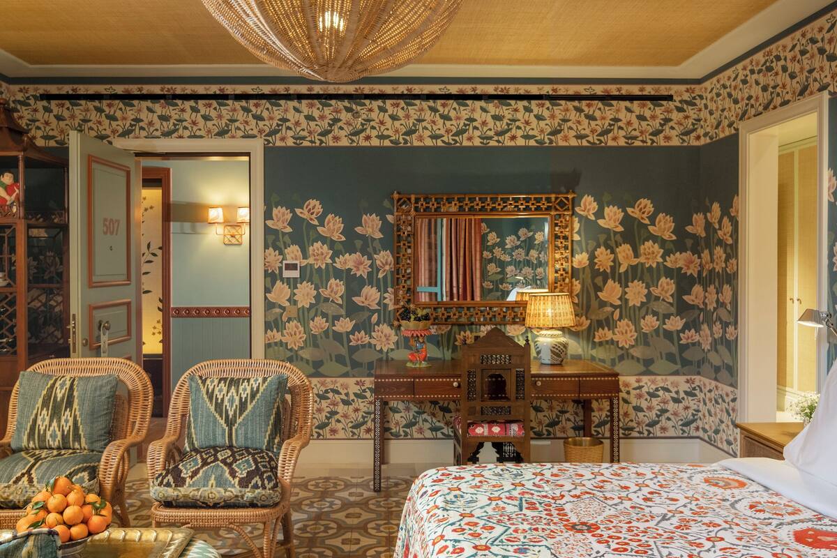 Une pièce avec un papier peint à motifs floraux, un lit recouvert d’une couvre-lit à motifs, des chaises en osier, un bureau en bois et un miroir.