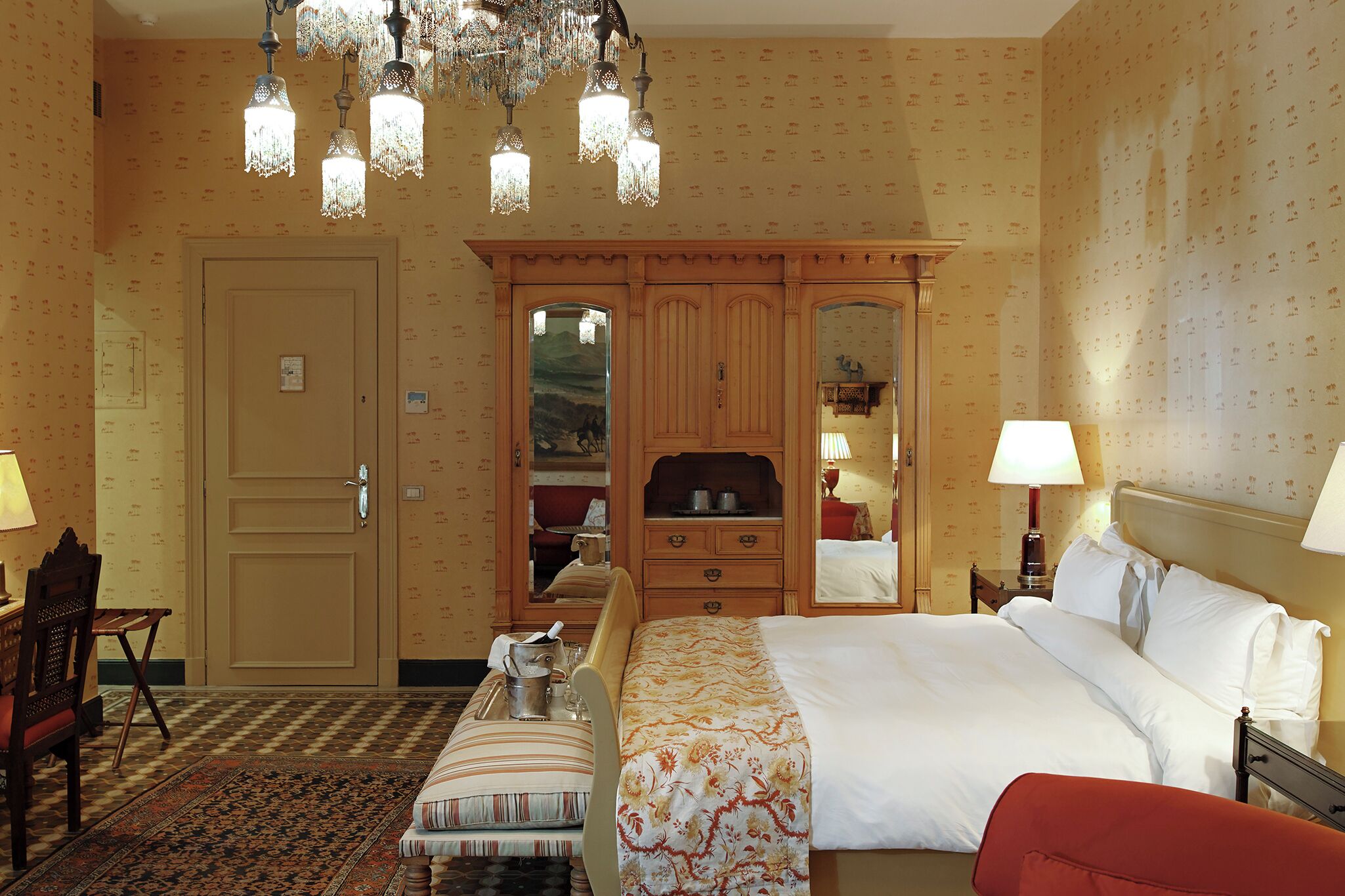 A hotel room with a bed, bedside tables, a wooden wardrobe, a desk with a chair, and a patterned rug.