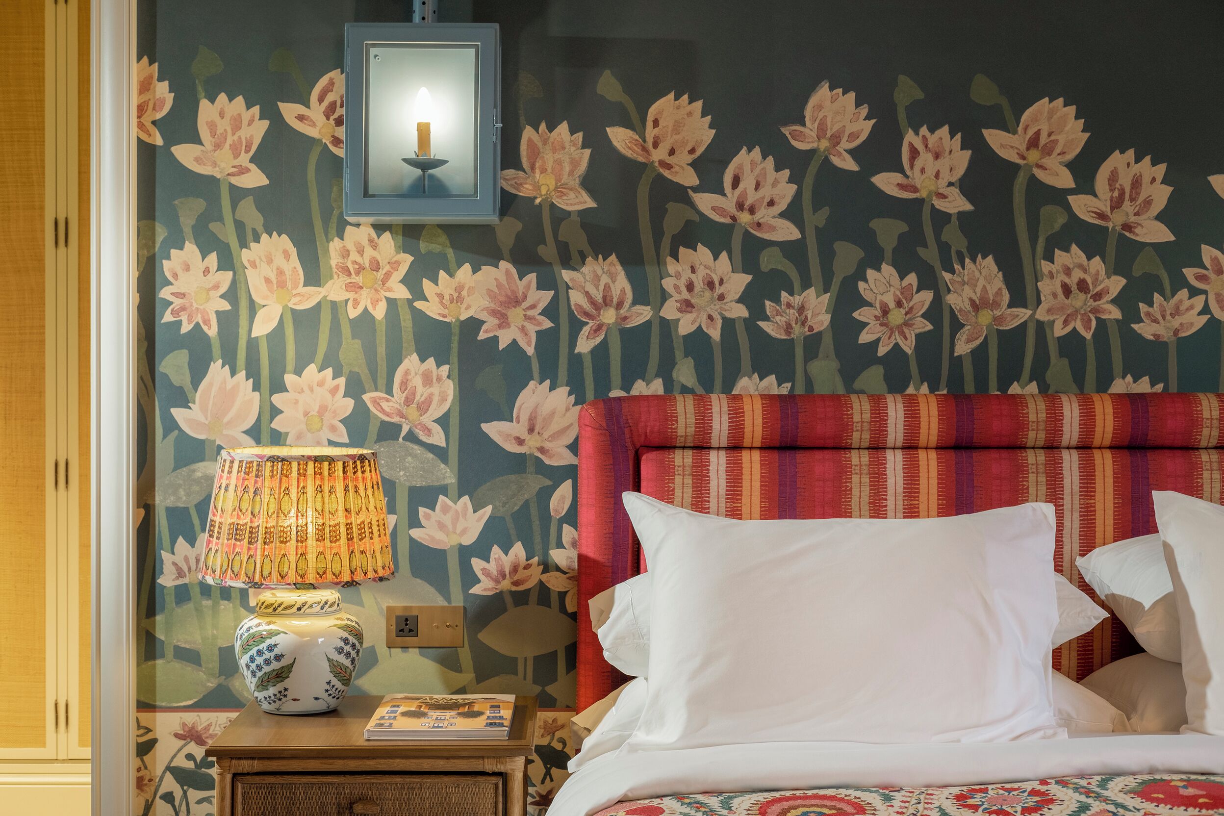 A bedroom with a floral wallpaper, a bedside lamp, and a bed with white pillows.
