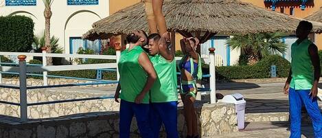 A group of people performing an acrobatic stunt in an outdoor setting with a building featuring blue windows and a thatched-roof structure in the background.
