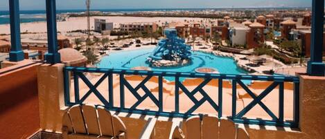 A balcony with two wooden chairs, a pool with a water slide, and a view of a beach and buildings.