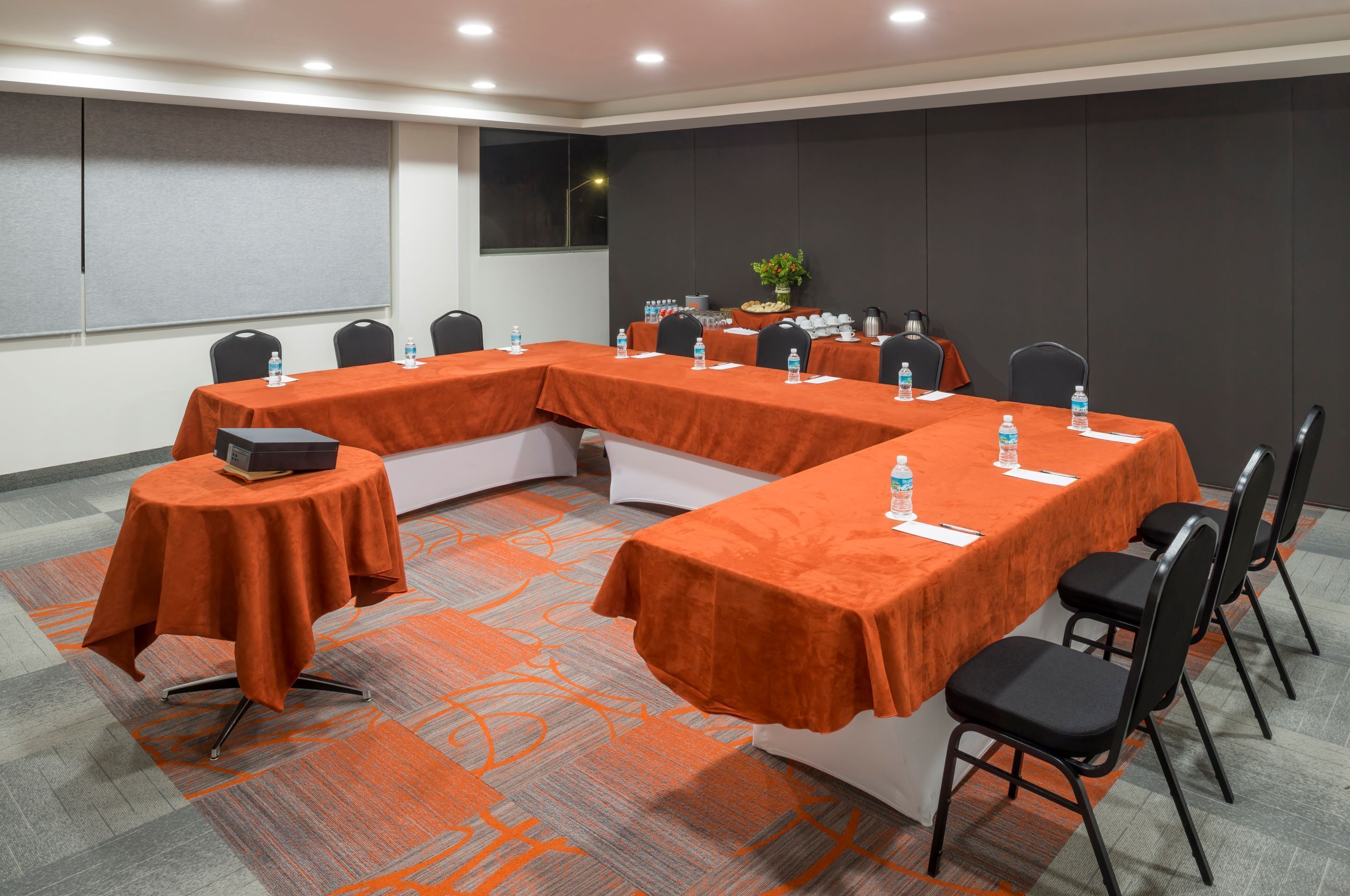 A conference room with a long table covered in orange cloth, surrounded by black chairs, and equipped with water bottles and a laptop.