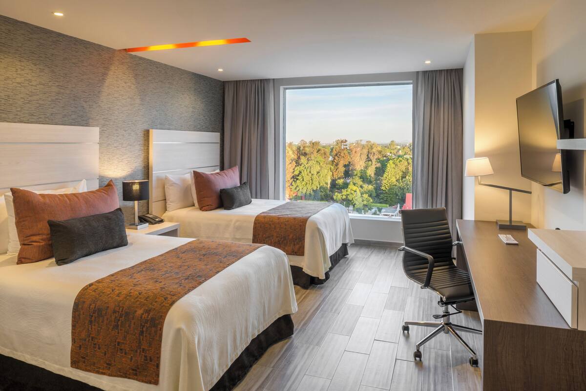 Une chambre d’hôtel avec deux lits, un bureau, une chaise, une télévision et une grande fenêtre donnant sur des arbres.