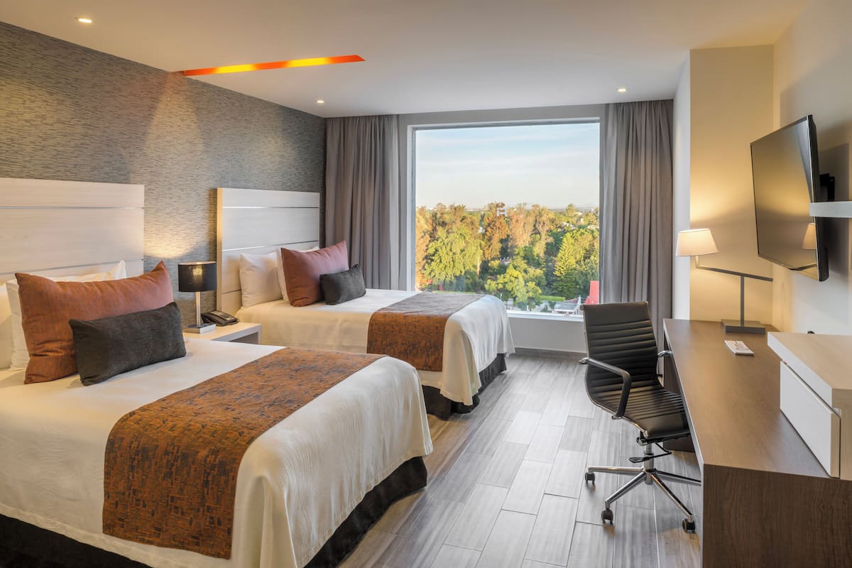 Ein Hotelzimmer mit zwei Betten, einem Schreibtisch, einem Stuhl, einem Fernseher und einem großen Fenster mit Blick auf Bäume.