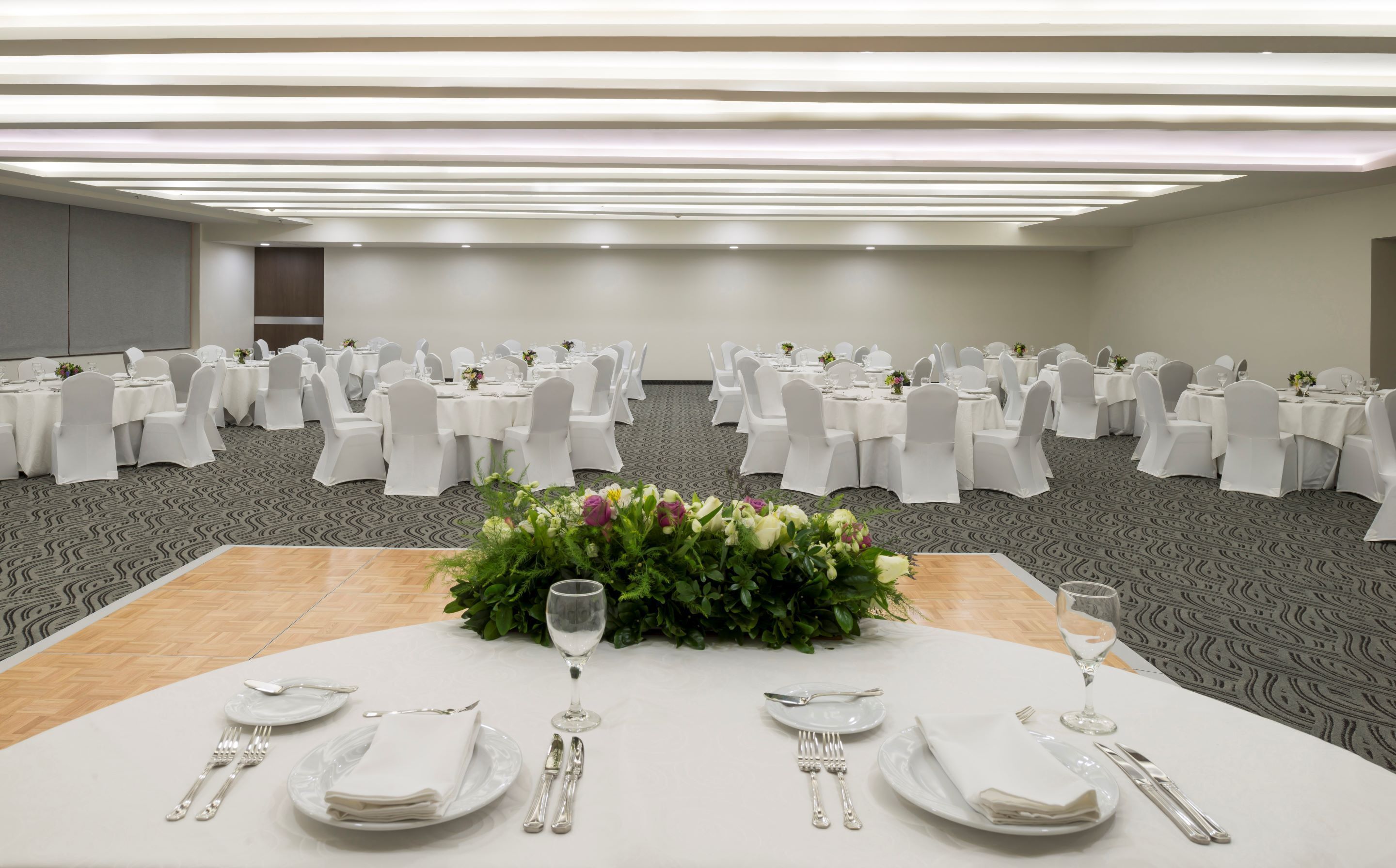 A banquet hall with round tables set for dining, white tablecloths, and chairs with white covers.