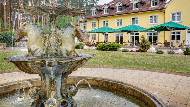 Ein Brunnen mit Pferdeskulpturen vor einem gelben Gebäude mit vielen Fenstern und grünen Sonnenschirmen.