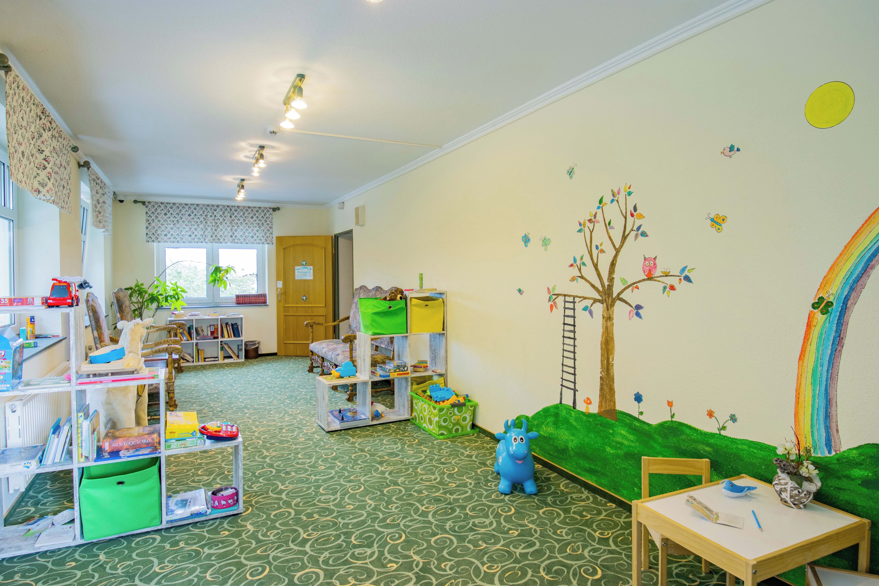 A children's playroom with a green carpet, shelves with toys, a tree mural, and a rainbow on the wall.
