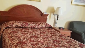 Une chambre d’hôtel avec un lit en bois, une couvre-lit à motifs floraux, deux tableaux encadrés au mur, une lampe de chevet et une chaise.