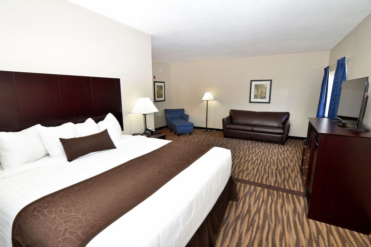 Ein Hotelzimmer mit einem großen Bett, einem braunen Kopfteil, einem blauen Sessel, einer braunen Couch, einem Schreibtisch mit einem Computer und einem Flachbildfernseher.
