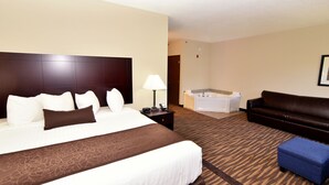 A hotel room with a large bed, a brown headboard, a white bedside table with a lamp, a blue ottoman, a brown leather sofa, and a separate bathroom with a bathtub.