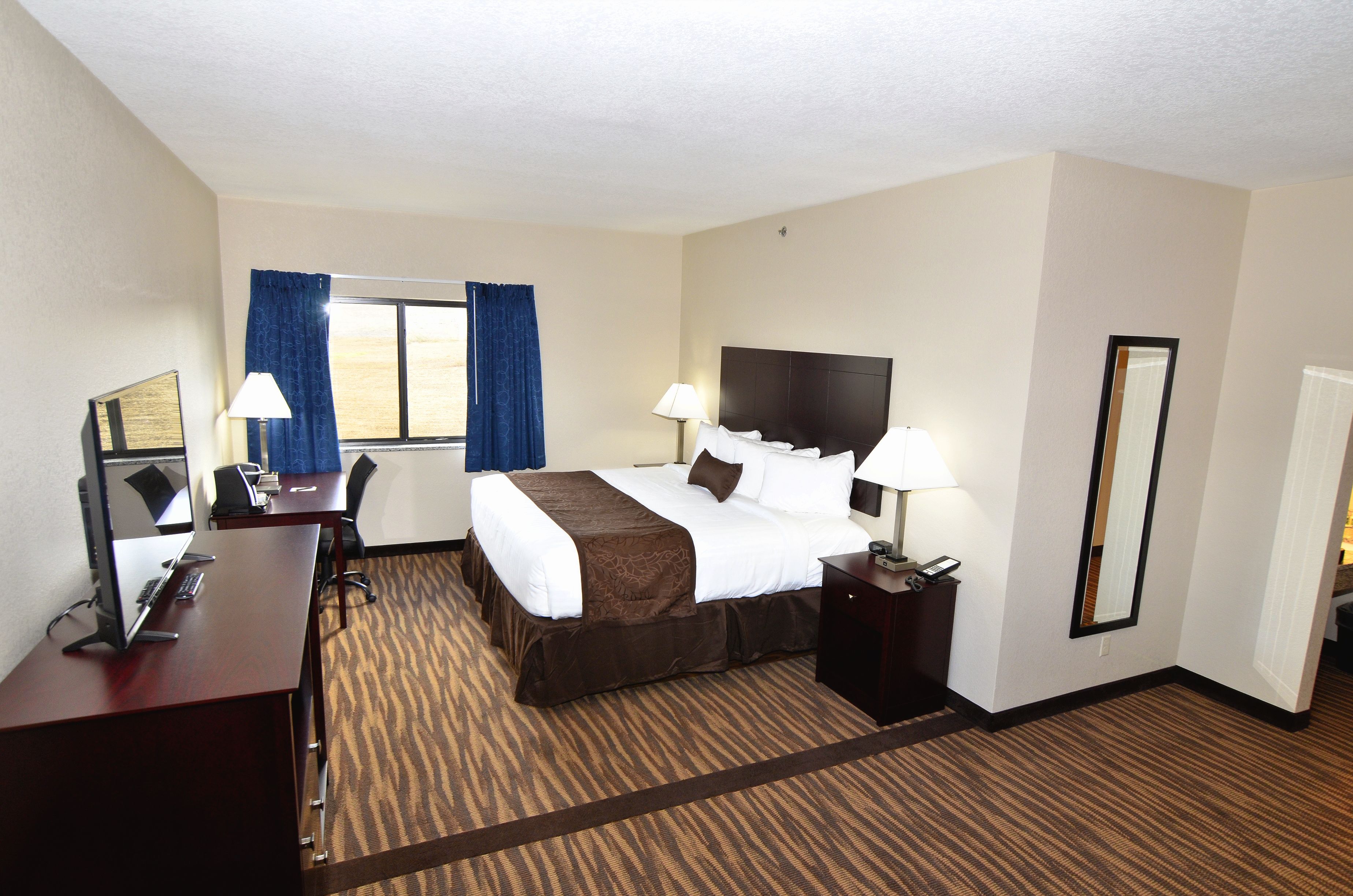 A hotel room with a large bed, a desk with a computer, a chair, a lamp, and a window with curtains.