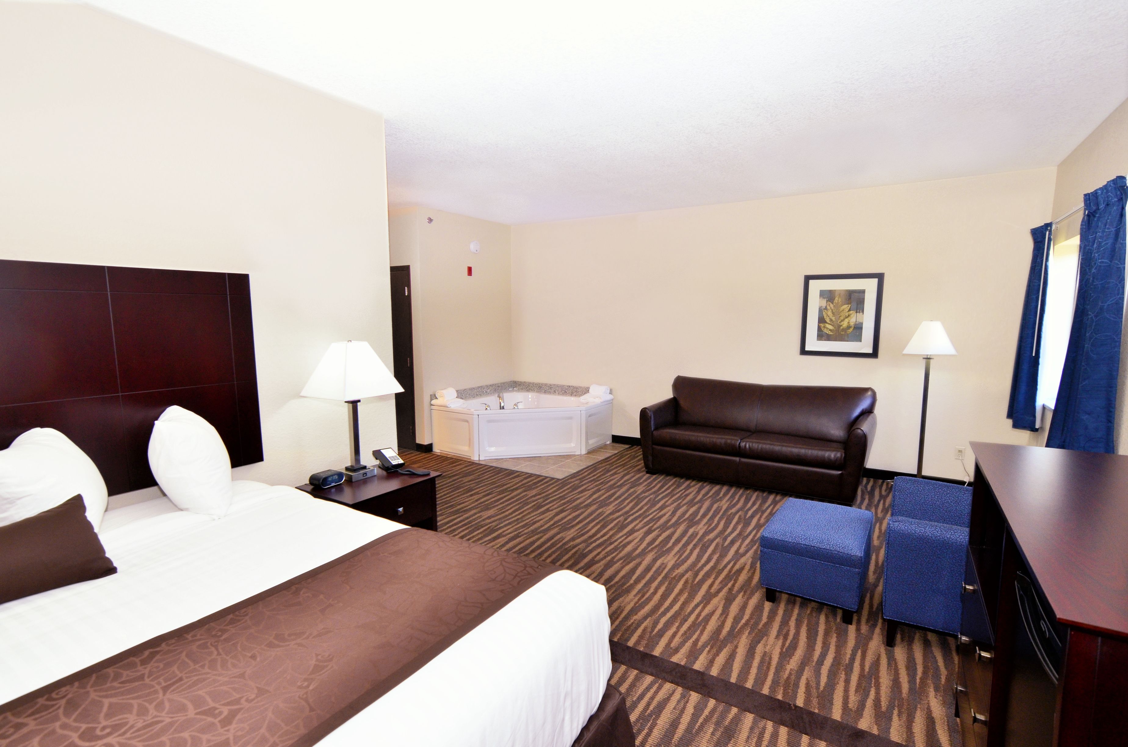 A hotel room with a large bed, a sofa, a small table, a desk, a chair, a lamp, a picture on the wall, and a bathtub.