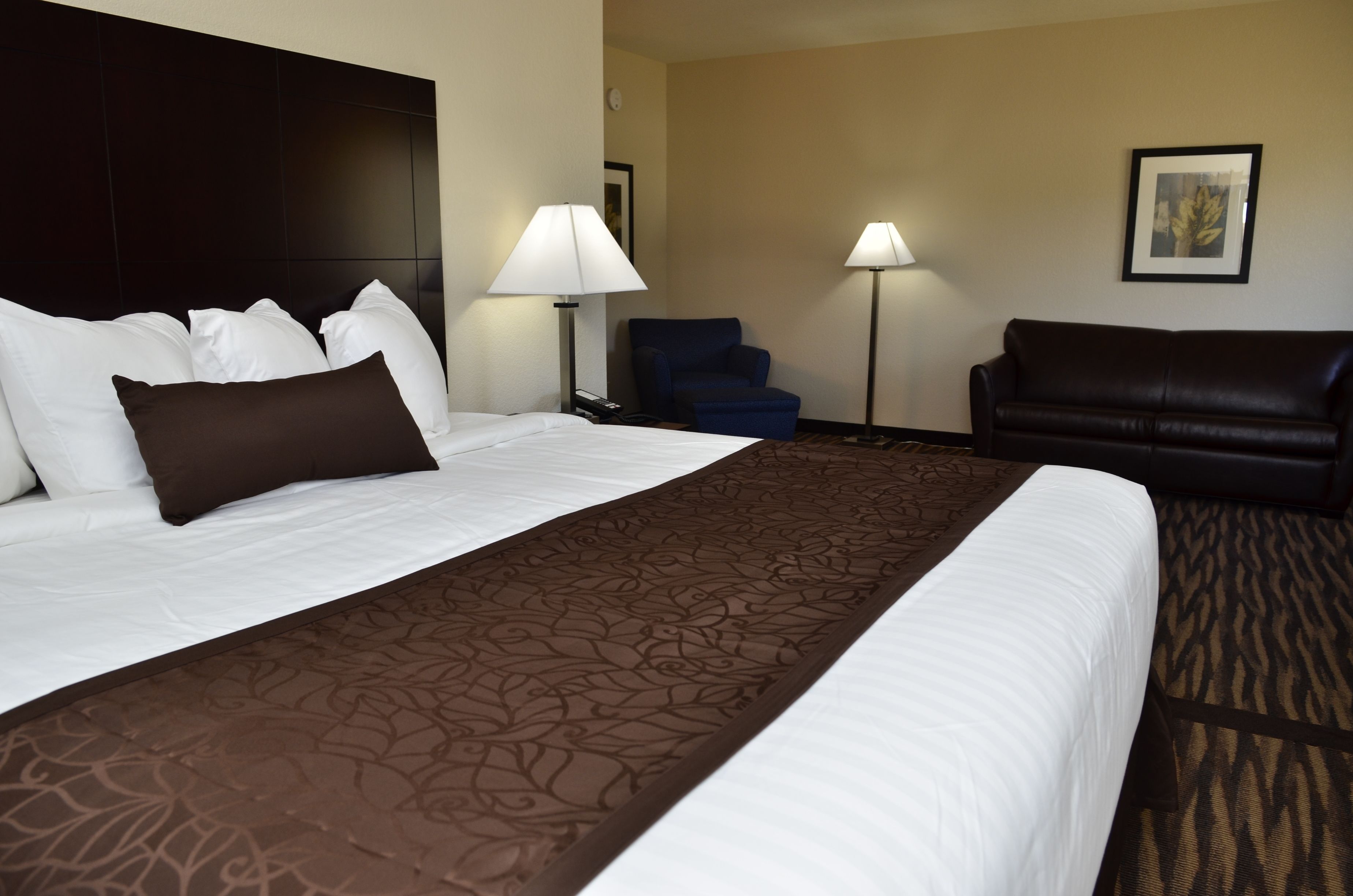 Quarto de hotel com cama grande, cabeceira escura, poltrona azul, sofá de couro preto e duas luminárias de chão.