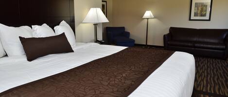 Quarto de hotel com cama grande, cabeceira escura, poltrona azul, sofá de couro preto e duas luminárias de chão.