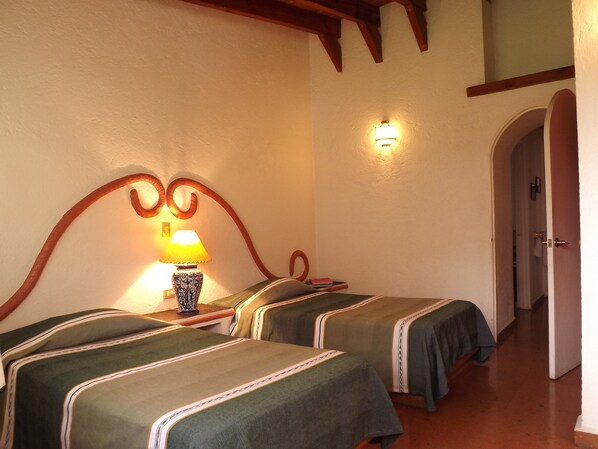 A room with two beds, each with a green bedspread and a decorative headboard. There is a table with a lamp between the beds, and a wall-mounted light fixture above the bed on the right.