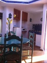 Un comedor con una mesa redonda y cuatro sillas, una cocina con armarios de madera y un microondas, y un reloj en la pared.
