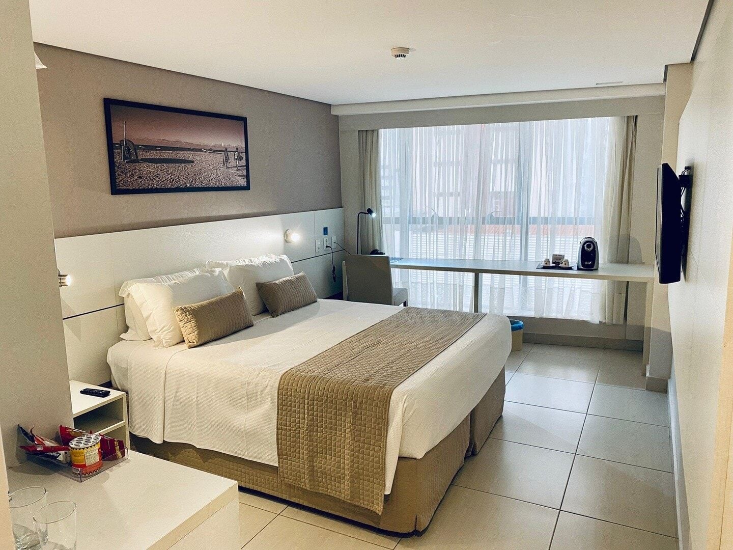 A modern hotel room with a large bed, a desk, a chair, a window with curtains, and a mounted television.