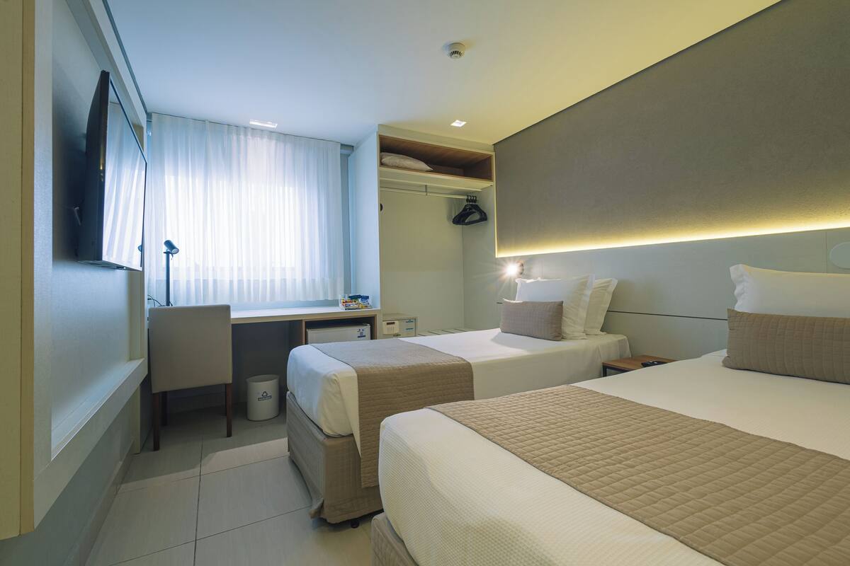 A hotel room with two beds, a desk, a chair, a TV, and a window with curtains.