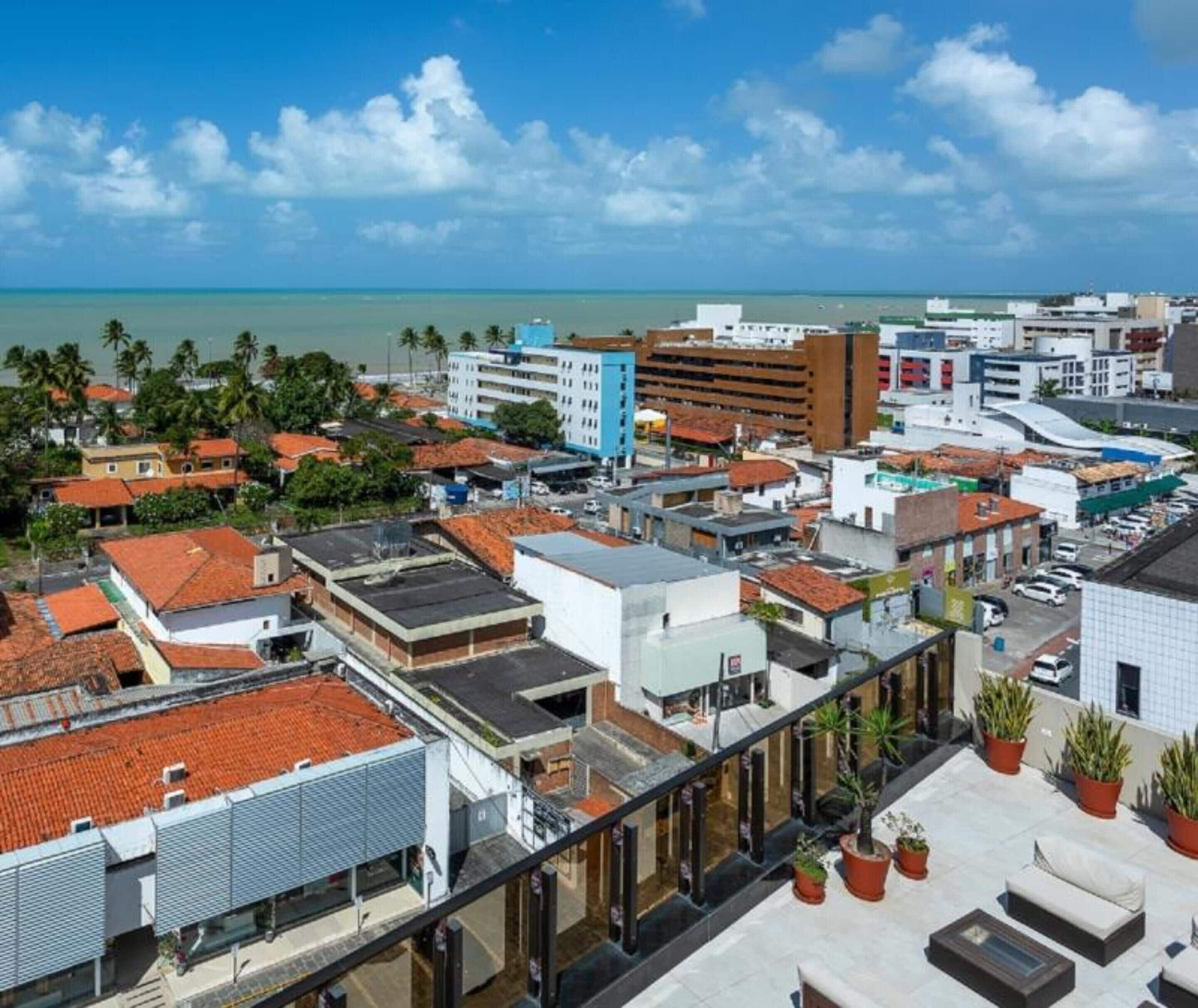 Un hôtel de grande hauteur, avec de nombreux étages, une terrasse sur le toit agrémentée de plantes en pot, et une vue sur l’océan et le paysage urbain.