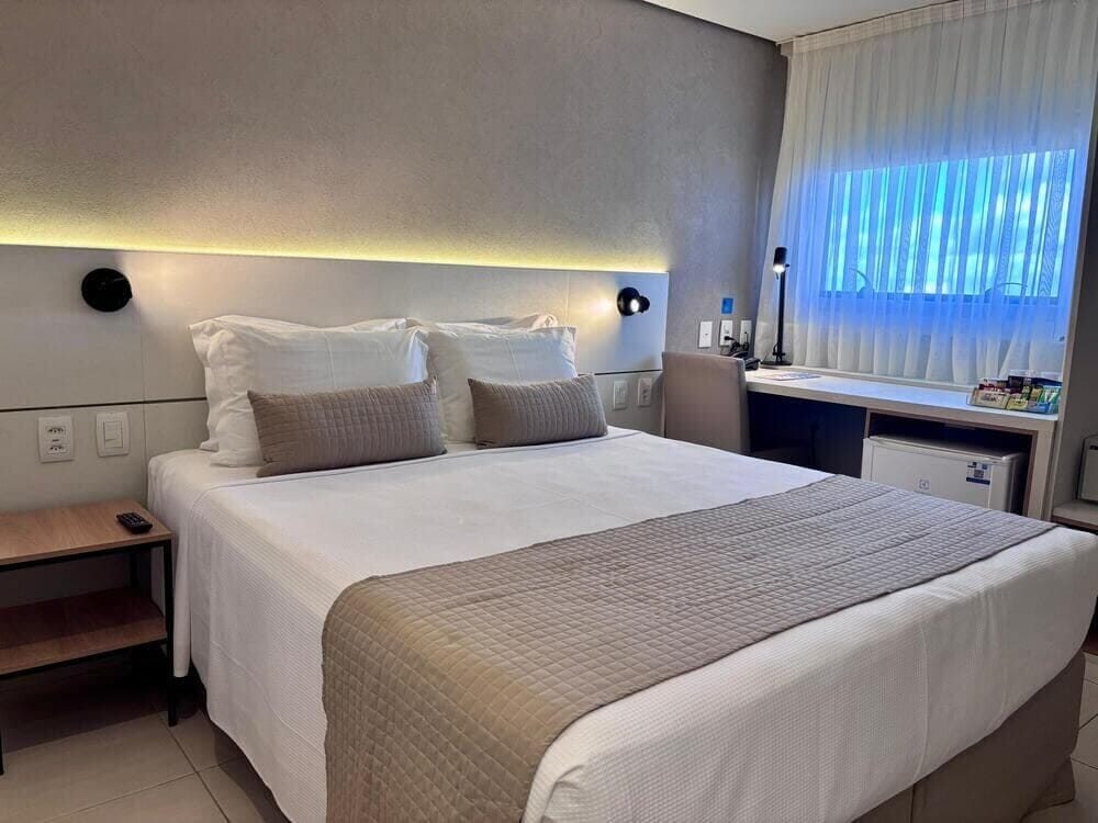 Quarto de hotel com cama, mesa de cabeceira, abajur e janela com cortinas.