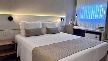 Quarto de hotel com cama, mesa de cabeceira, abajur e janela com cortinas.