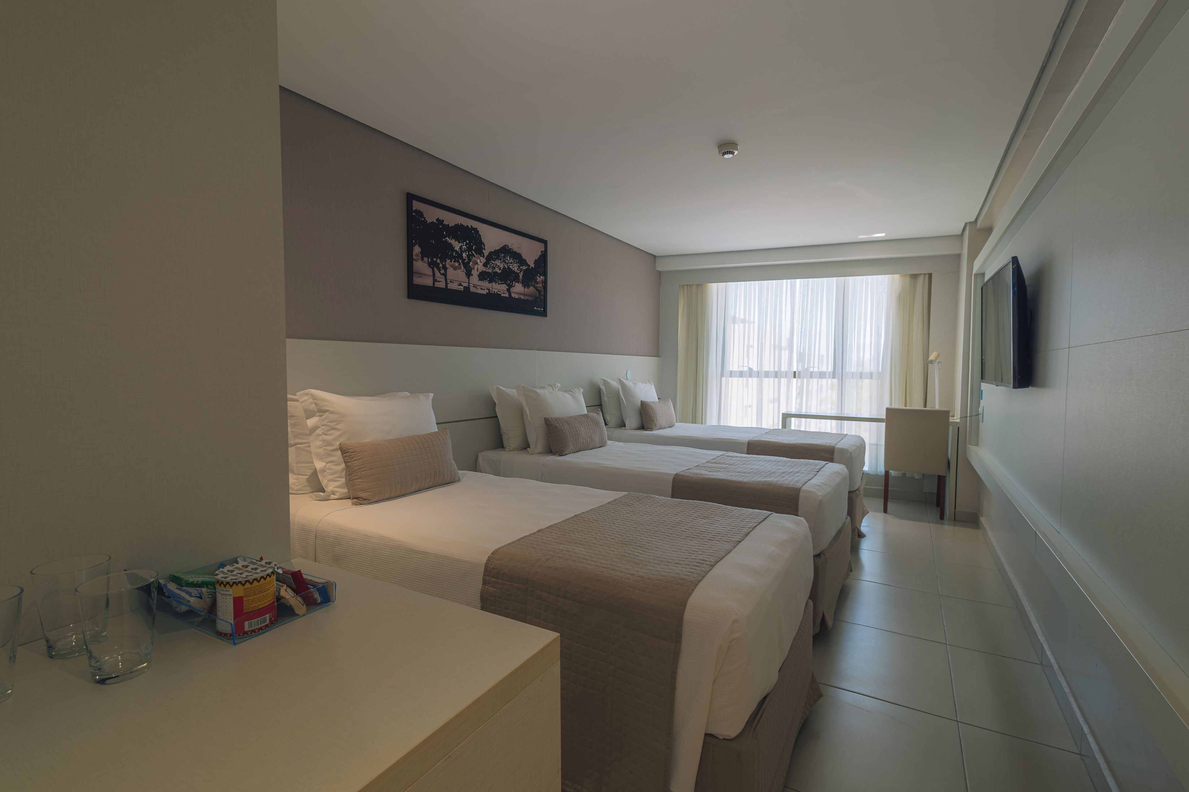 Habitación de hotel con dos camas, televisión, ventana con cortinas y mesita de noche con vasos y una caja.