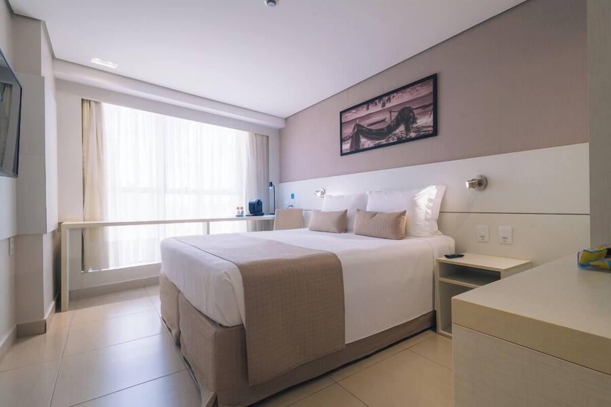 A modern hotel room with a large bed, bedside tables, a desk, and a window with curtains.