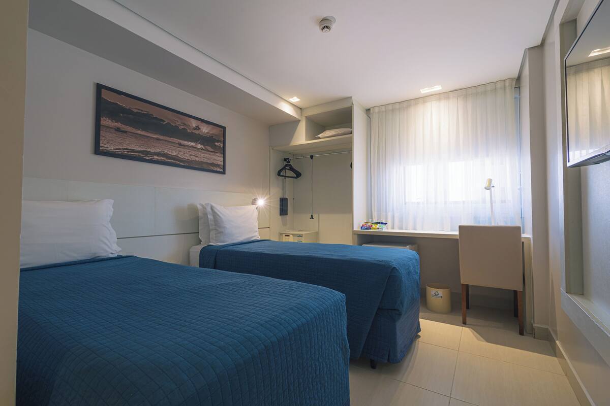 A hotel room with two beds, a desk, a chair, a wardrobe, and a window with curtains.