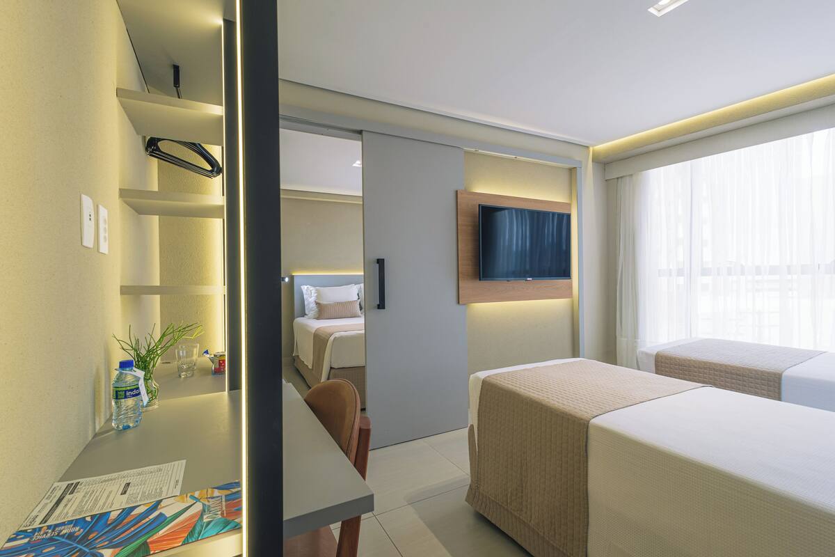 A hotel room with a bed, a TV mounted on the wall, a small table with a water bottle, and a window with curtains.