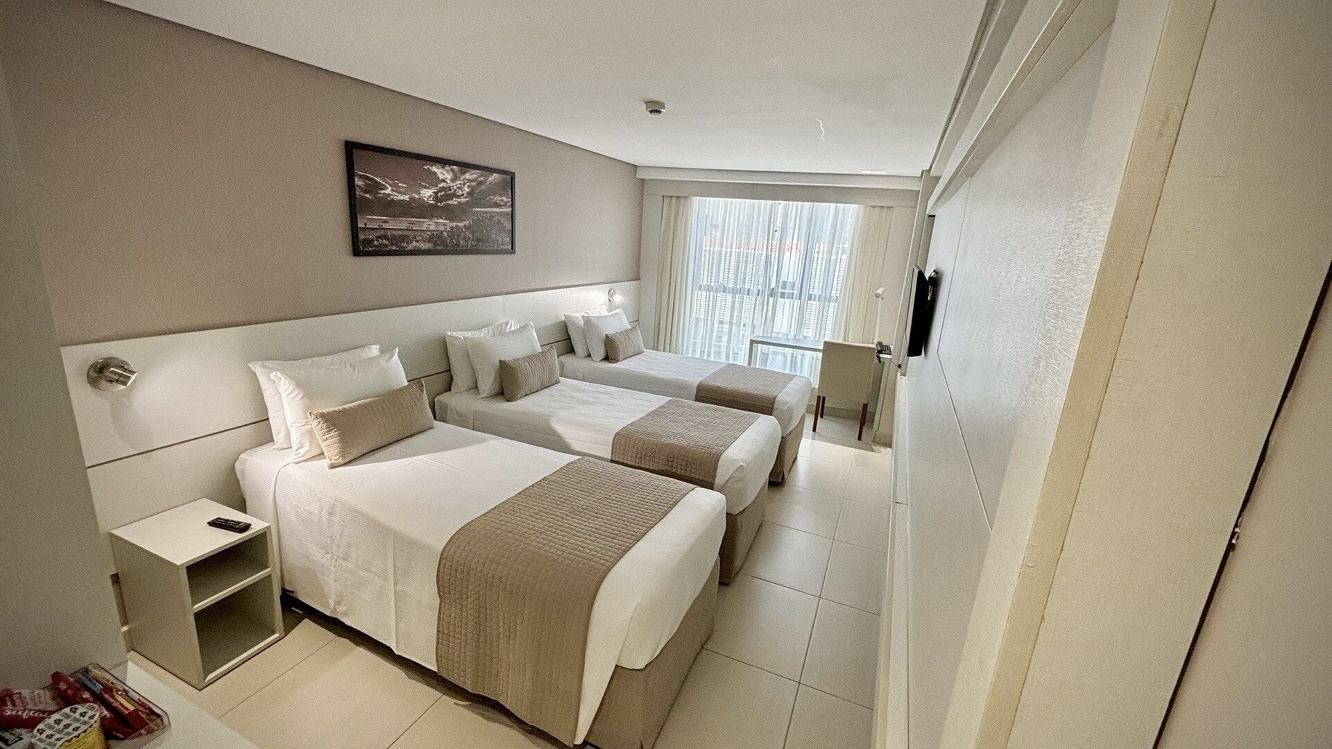 A hotel room with two beds, a bedside table, a small desk, and a window with blinds.