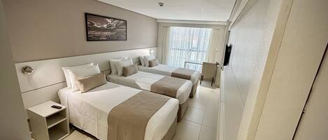 A hotel room with two beds, a bedside table, a small desk, and a window with blinds.
