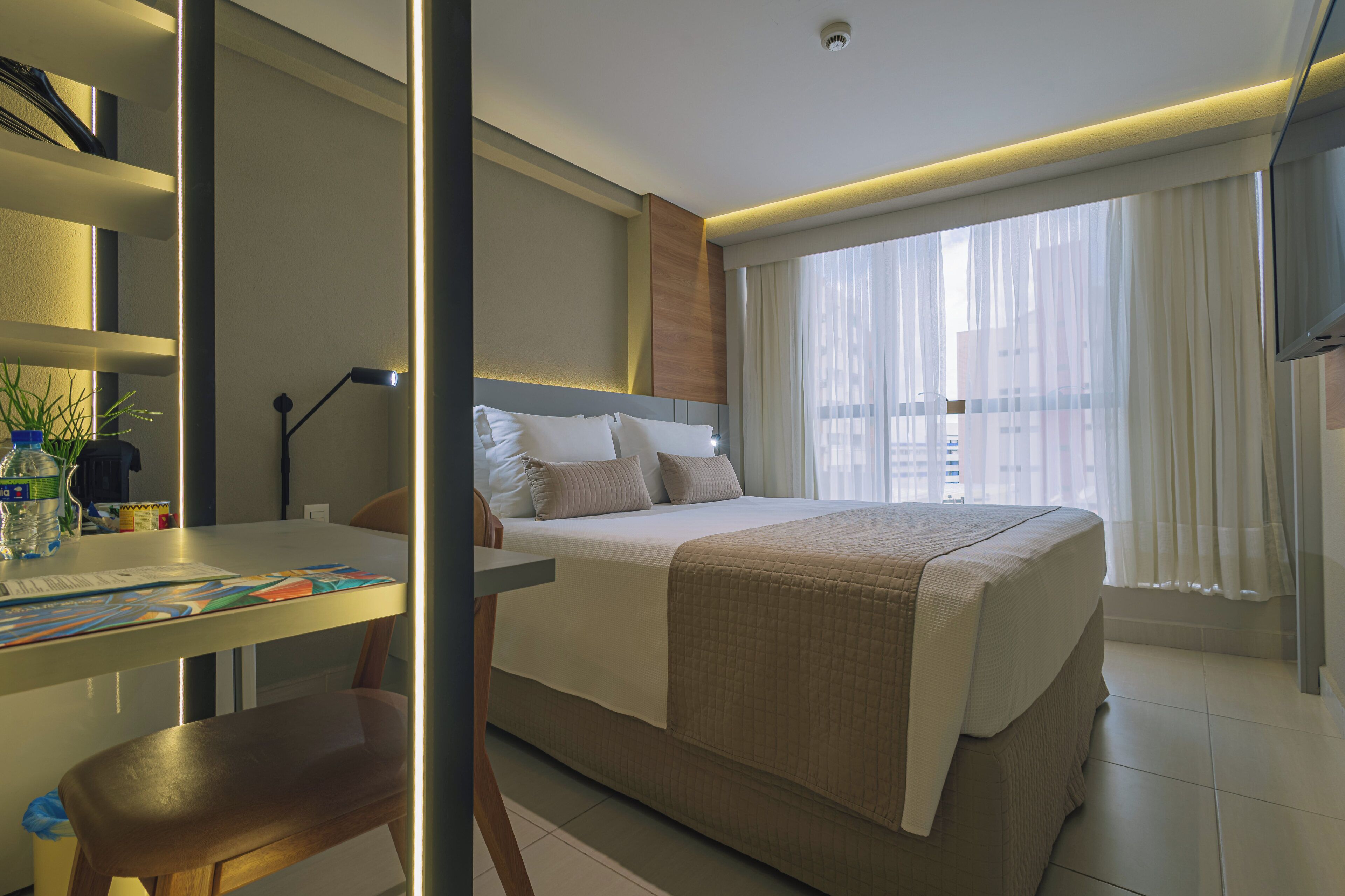 A modern hotel room with a neatly made bed, a desk with a chair, and a large window with curtains.