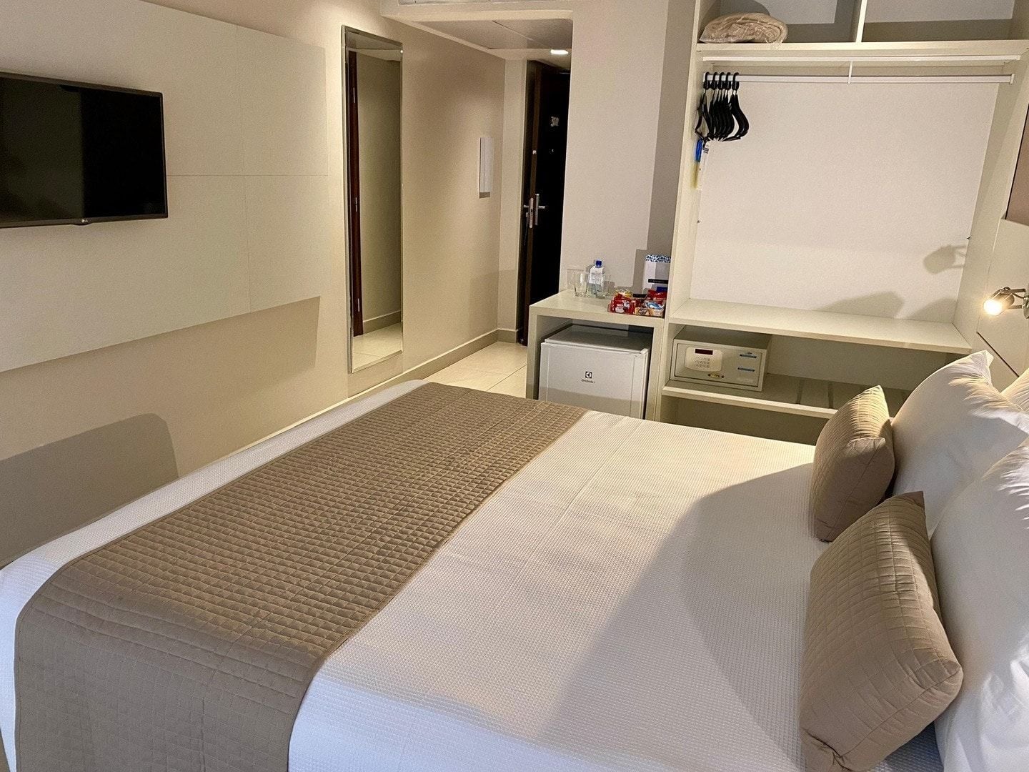 A hotel room with a bed, a bedside table, a TV, a wardrobe, and a small refrigerator.