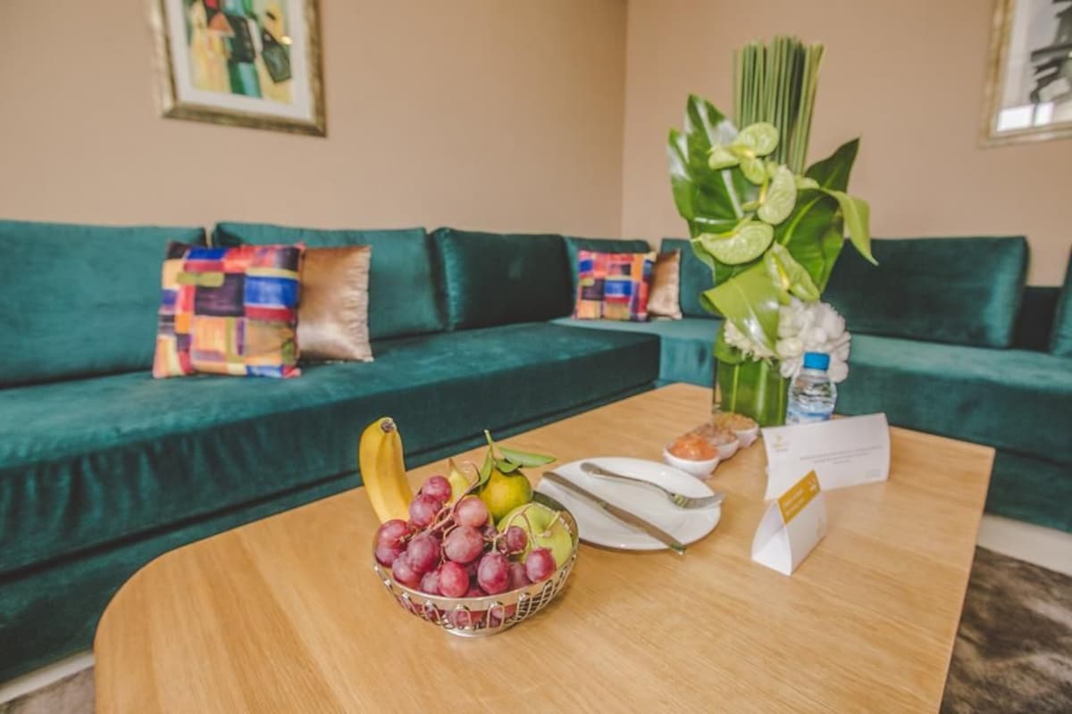 A room with a green sofa, a wooden coffee table with fruits, a vase with flowers, and a framed picture on the wall.