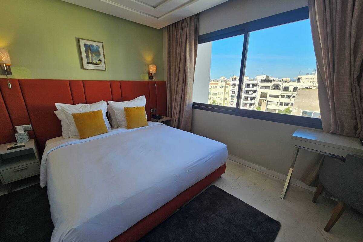 Ein Hotelzimmer mit einem großen Bett, einem Schreibtisch, einem Nachttisch, einem Bild an der Wand und Blick auf Gebäude durch das Fenster.