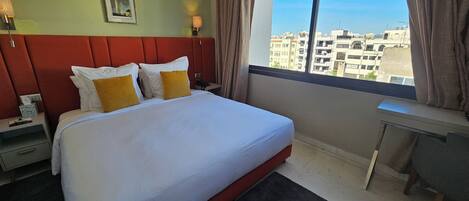 A hotel room with a large bed, a desk, a nightstand, a picture on the wall, and a view of buildings through the window.