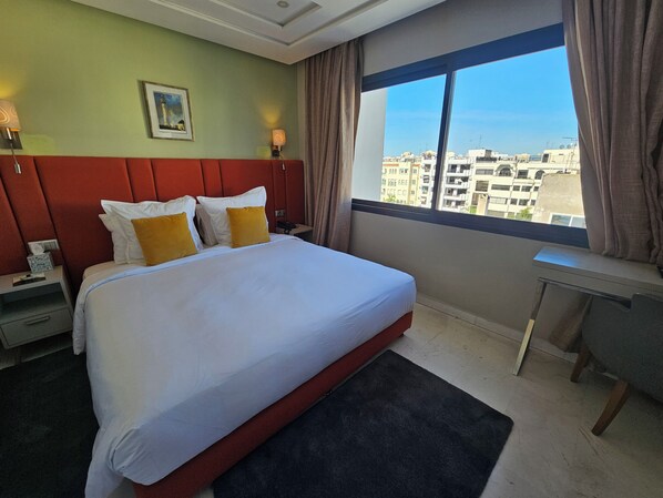 Habitación de hotel con una cama grande, un escritorio, una mesita de noche, un cuadro en la pared y vistas a edificios a través de la ventana.