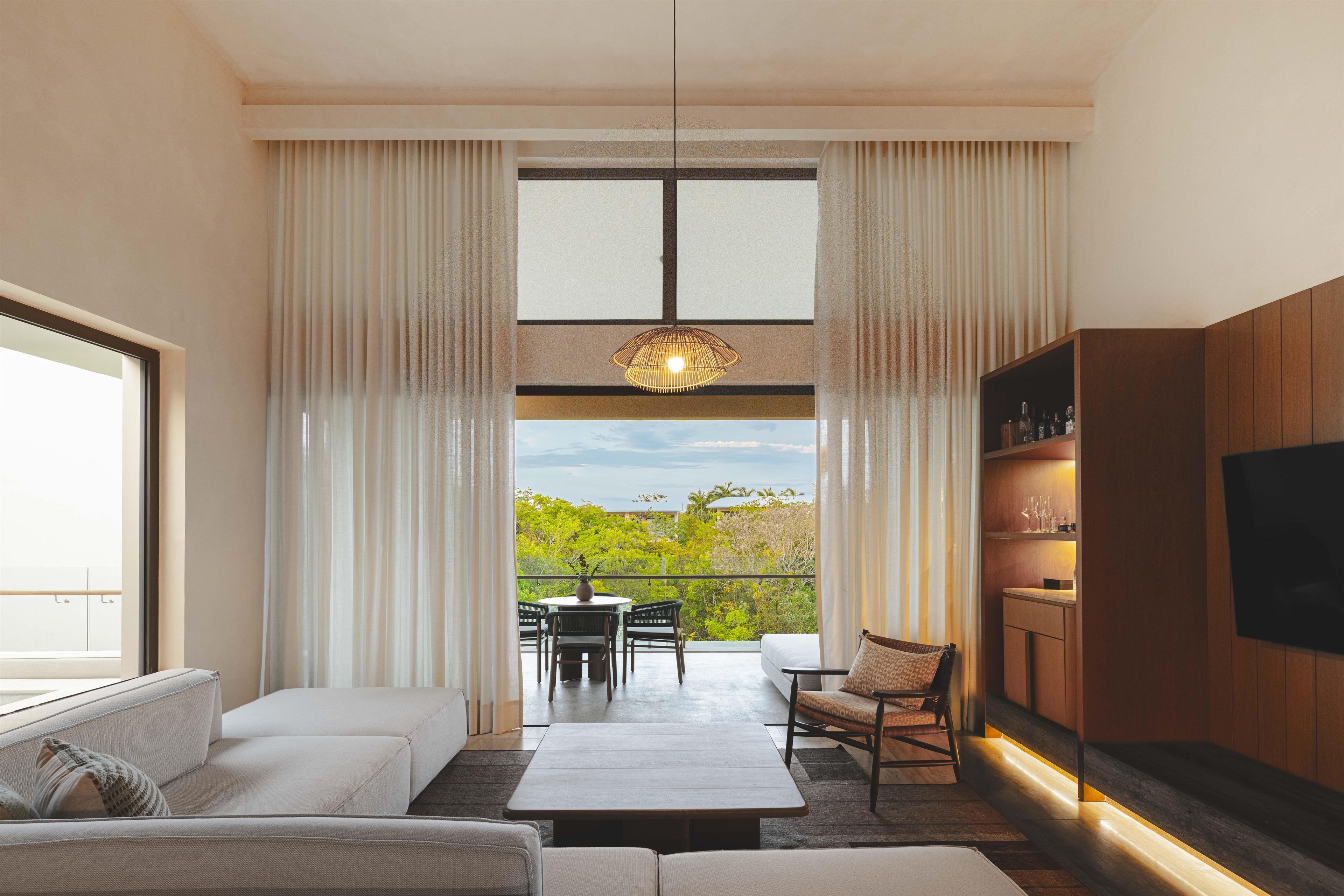 A modern living room with a sofa, a dining table, and chairs, a TV mounted on the wall, and a balcony with a view of greenery and the sky.