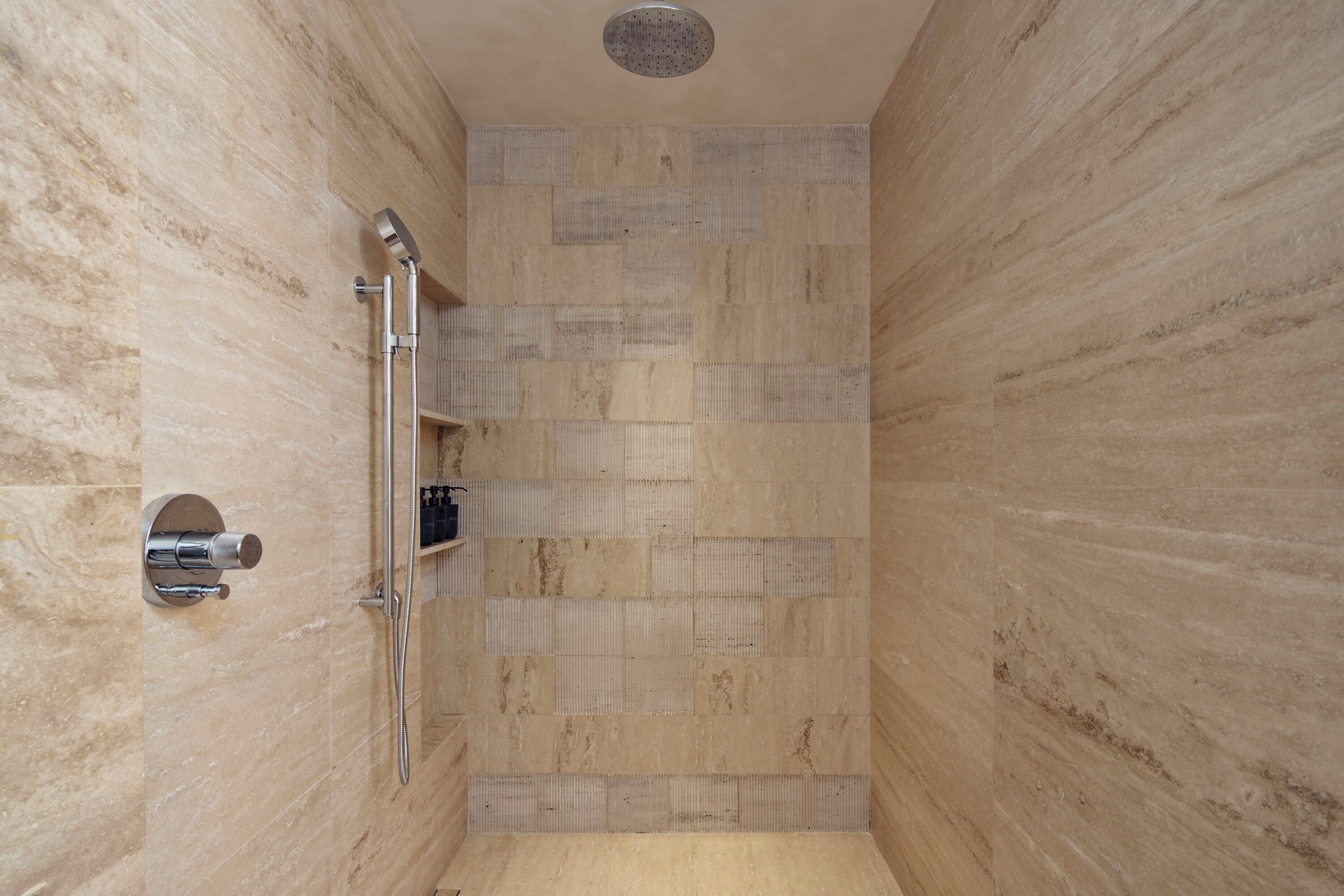 Une douche avec un robinet mural et une pomme de douche à main, une petite étagère pour les articles de toilette et un sol carrelé.