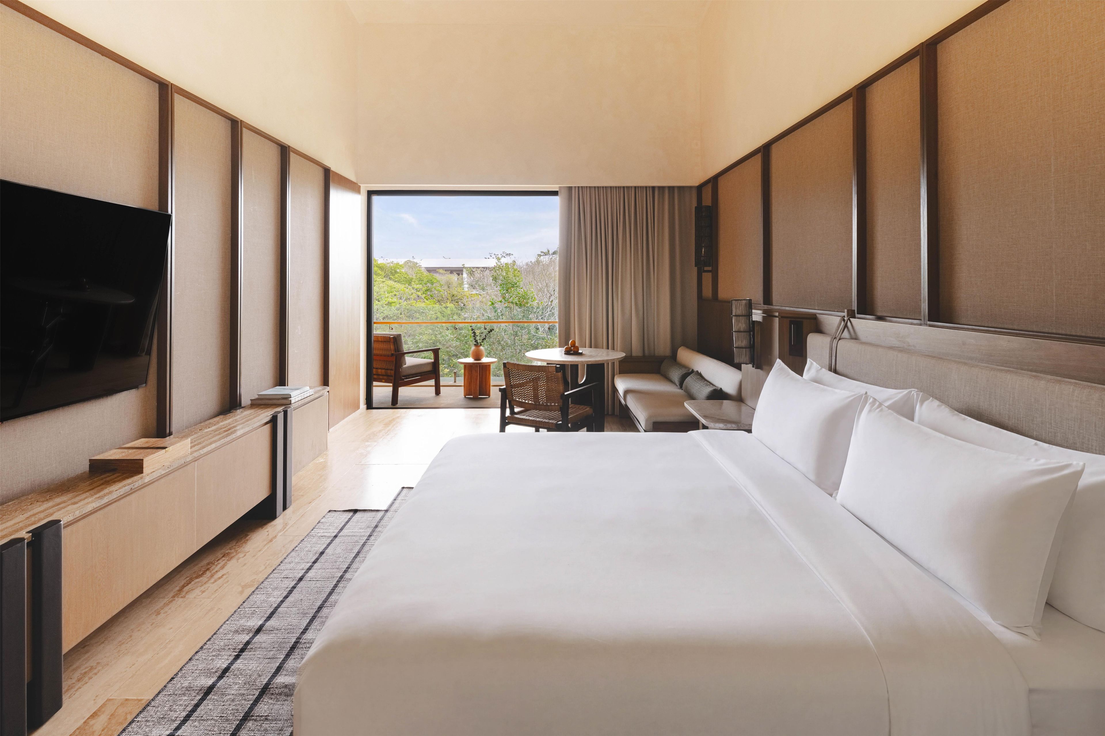 A hotel room with a large bed, a TV, a balcony with seating, and a view of greenery.