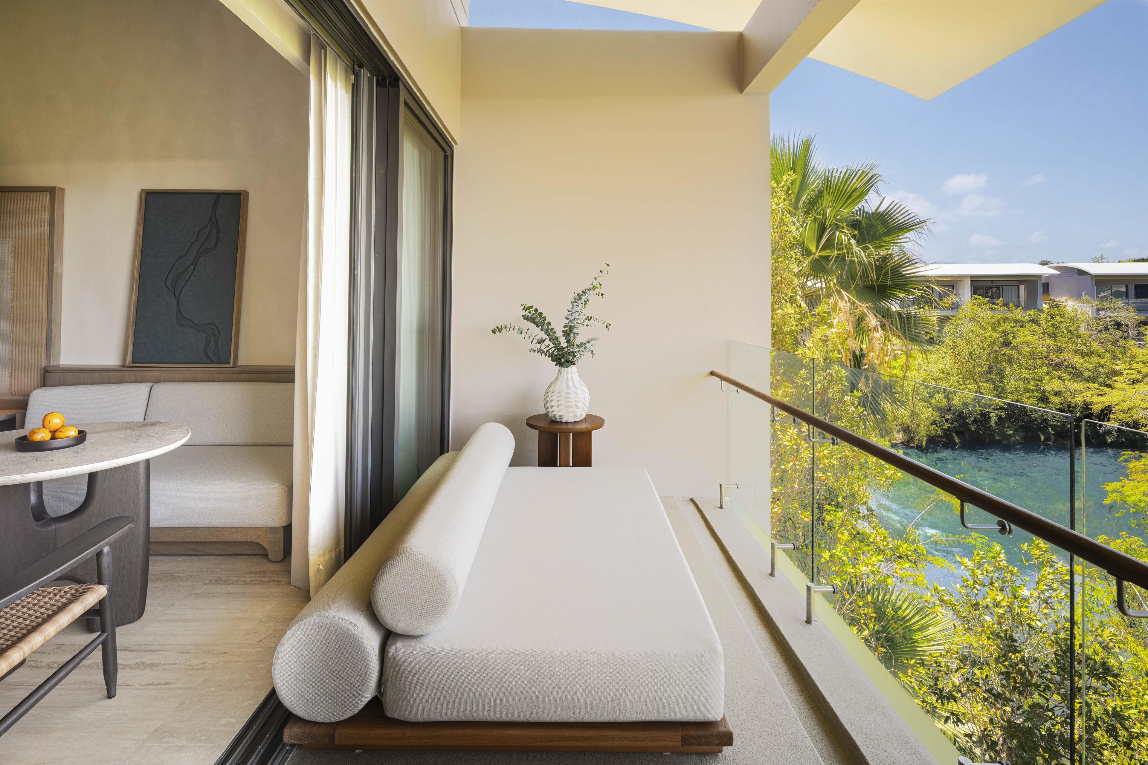 Un balcon avec un lit, une petite table avec des fruits, un vase avec des plantes et une vue sur une rivière et la verdure.