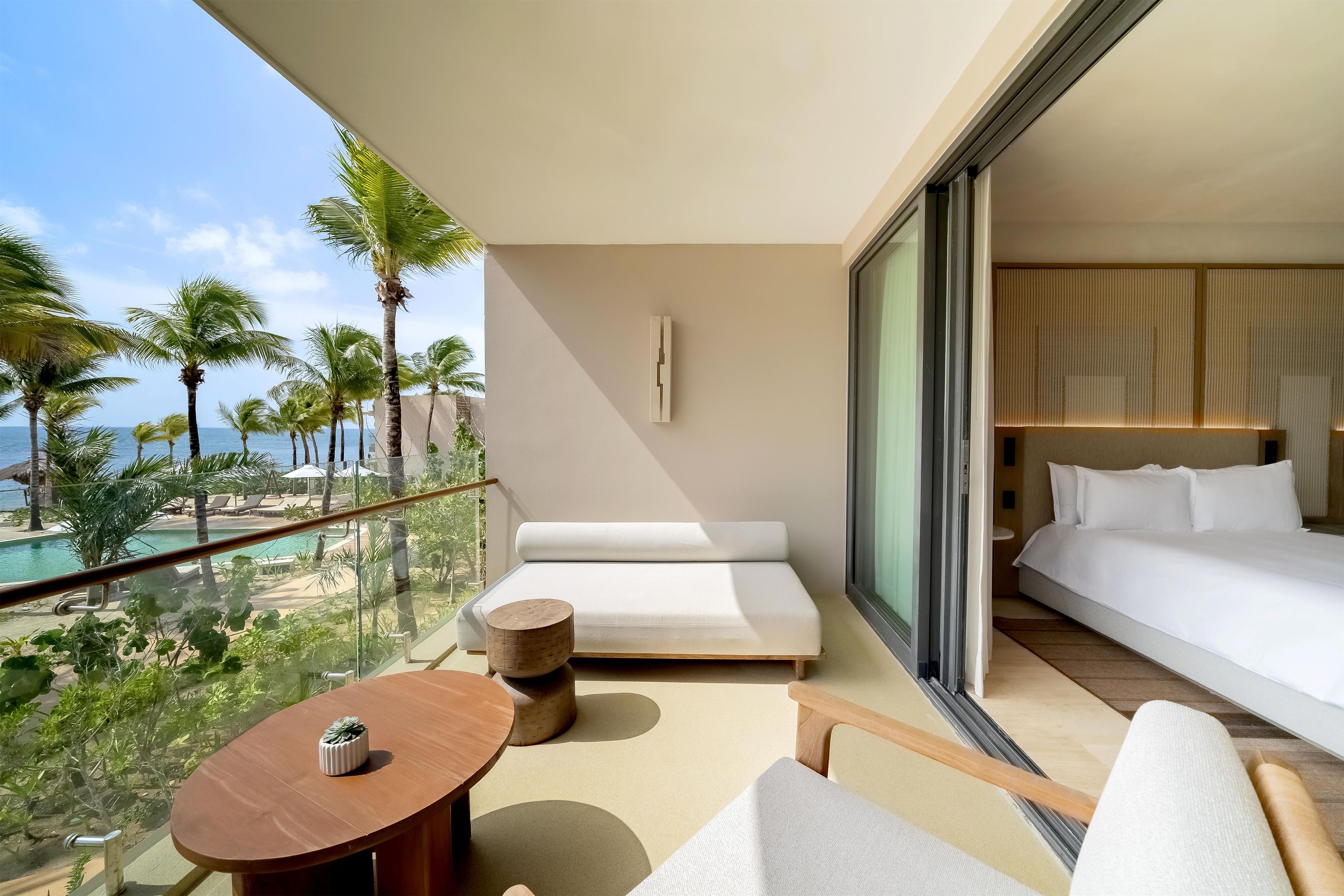 A balcony with a bed, a small table, and a view of a pool and palm trees.