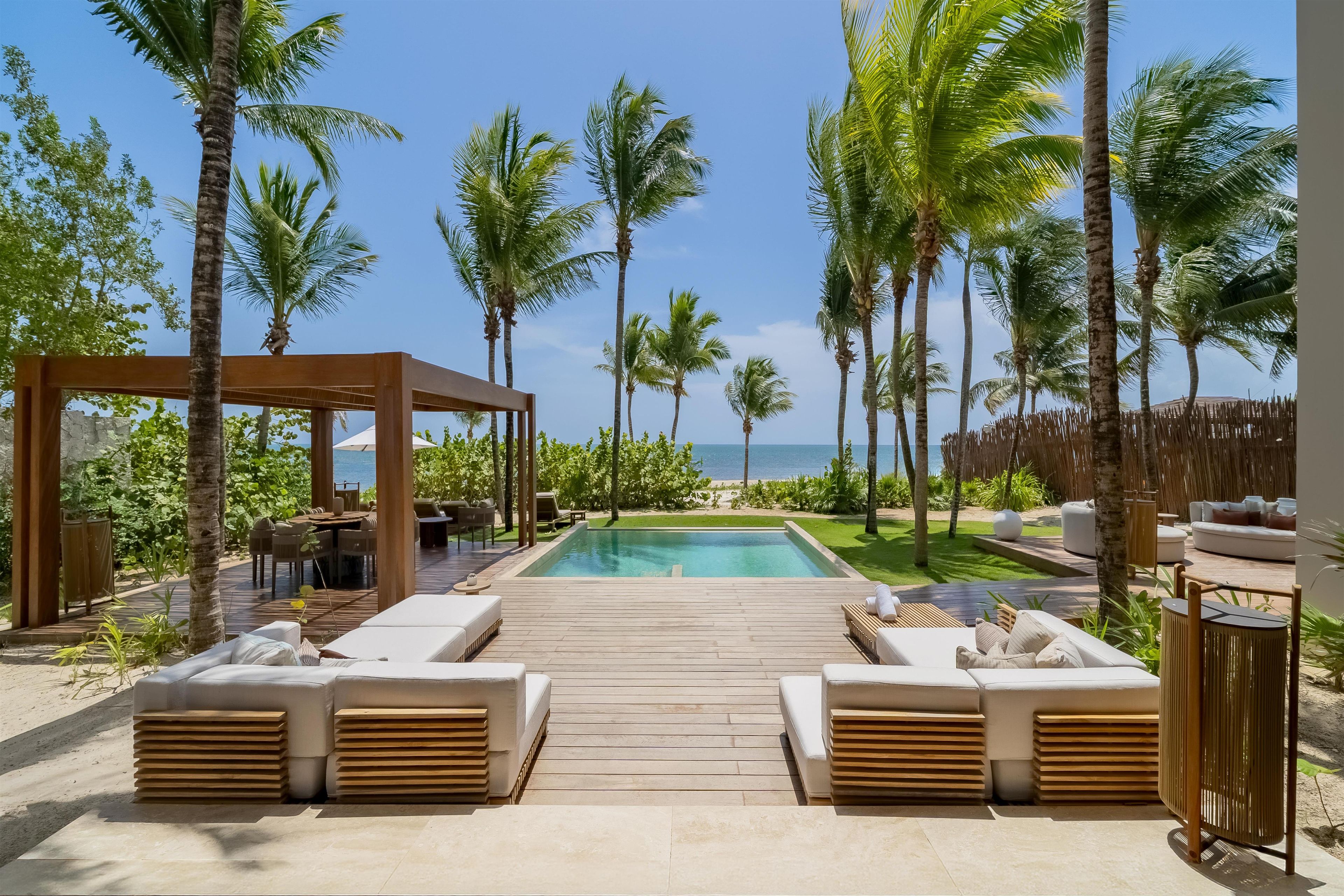 Zona de piscina con sillones de descanso y terraza de madera, rodeada de palmeras y vista al océano.
