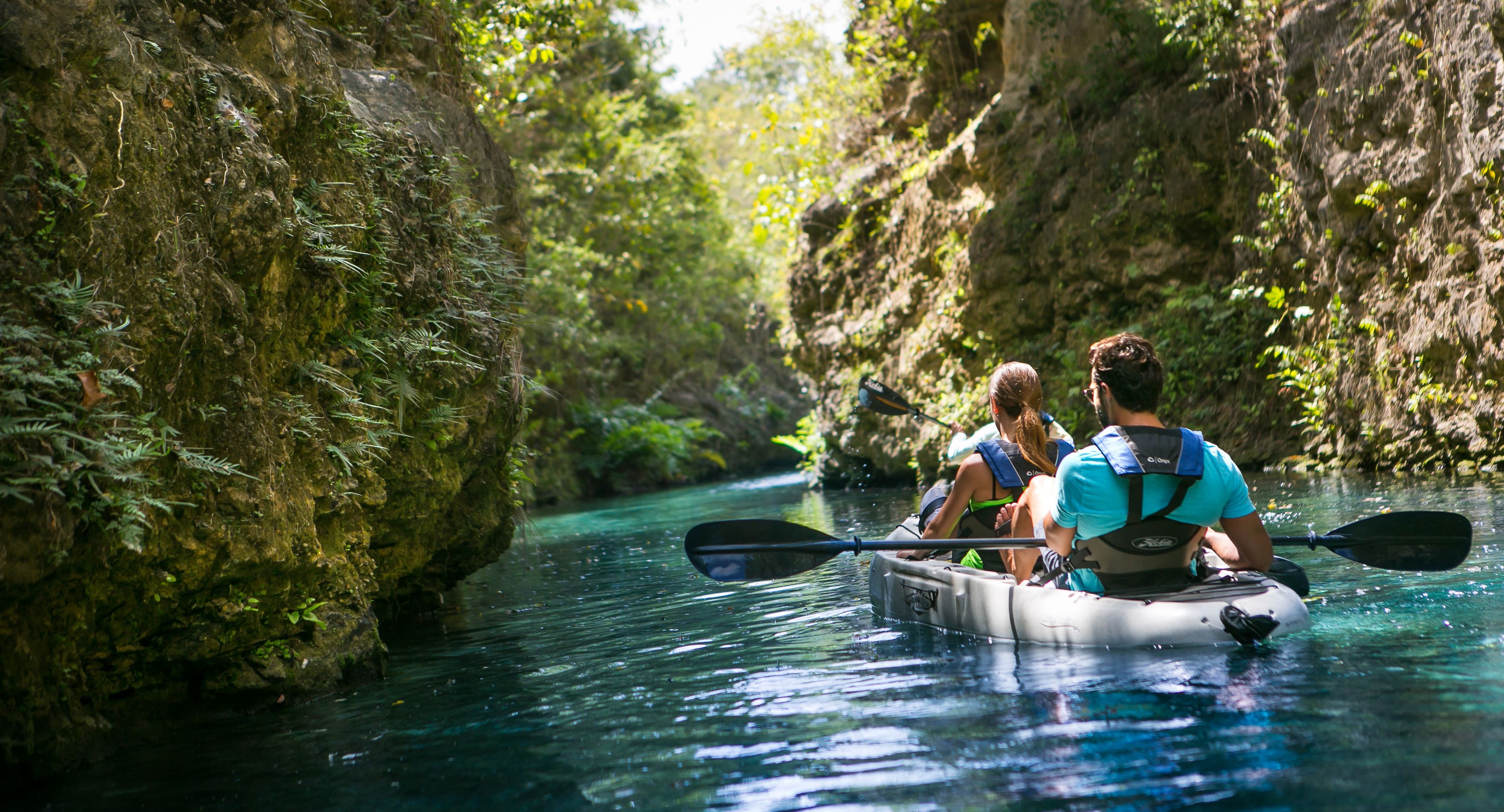 Una pareja practica kayak en un río estrecho y rocoso, rodeada de exuberante vegetación.