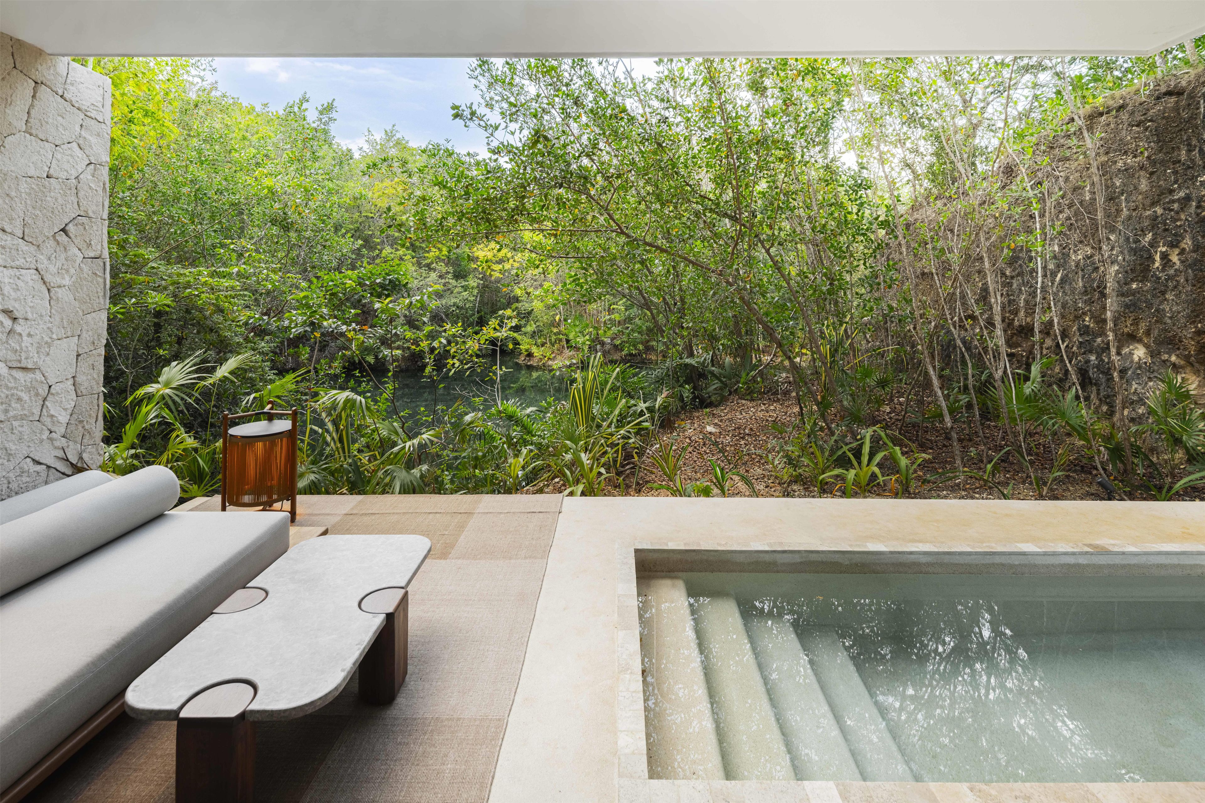Un espacio exterior moderno con zona de estar, pared de piedra, piscina y abundante vegetación.